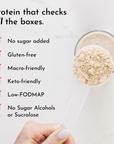 A hand holds a measuring scoop filled with beige protein powder against a blurred background of a drink.