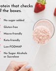 A hand holds a clear scoop of pink protein powder above a glass of blended drink, with strawberries in the background.