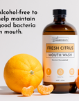 Brown glass bottle of Fresh Citrus mouthwash with whole and sliced oranges beside it on a light background.
