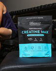 Black and cyan pouch of Creatine Max supplement sitting on exercise bench with muscular person in navy tank top beside it.