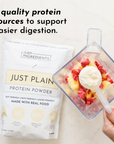 White protein powder in measuring scoop above blender with berries and fruit; product package displayed beside.