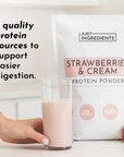 A hand holds a glass of pink protein shake next to a bag of Strawberries & Cream Protein Powder on a countertop.