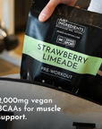 Black pouch labeled "Strawberry Limeade Pre-Workout" with text about 2,000mg vegan BCAAs, held above a gym bag.