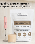 White pouch of Snickerdoodle Protein Powder with pink milk frother and glass of tan-colored beverage.