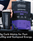 Black pouch of grape-flavored endurance drink with 35g carbohydrates and 500mg sodium, held next to matching black tumbler.