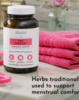 Dark glass bottle labeled "PMS Support Hormone Support" with white cap next to pink folded towels and accessories.