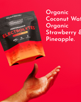 Black and orange electrolyte packets with strawberry tropic flavor labeling above an open hand on red background.