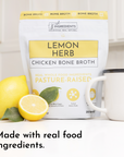 White pouch of lemon herb chicken bone broth with a lemon and green leaves beside a white mug on a countertop.