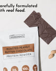 White pouch of roasted peanut butter chocolate protein powder next to chocolate bar and peanuts on a marble surface.