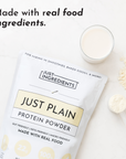 White protein powder package labeled "Just Plain" next to glass of mixed powder and measuring spoon on white surface.