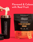 Orange recovery drink powder in black pouch next to black shaker bottle with powder being poured on red background.