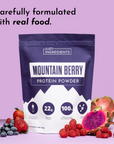 Purple packaging of Mountain Berry Protein Powder with berries and a dragon fruit on a pink background.