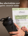 A person holds a shaker bottle and a package of Strawberry Limeade Electrolytes featuring key ingredients.