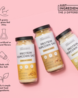 Three jars of Protein Popcorn Salt in Cheddar Cheese, Caramel Corn, and Parmesan Cheese flavors on a pink background.