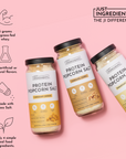 Three jars of protein popcorn salt in cheddar cheese, caramel corn, and parmesan cheese flavors on a pink background.