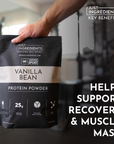 Black and white bag of Vanilla Bean Protein Powder held in a hand, with gym equipment blurred in the background.