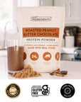 Hand holding a glass of chocolate protein shake next to a bag of Roasted Peanut Butter Chocolate Protein Powder and shelled peanuts.