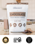 Hand holding a clear scoop of beige protein powder above a glass with a chocolate shake and chocolate pieces behind.
