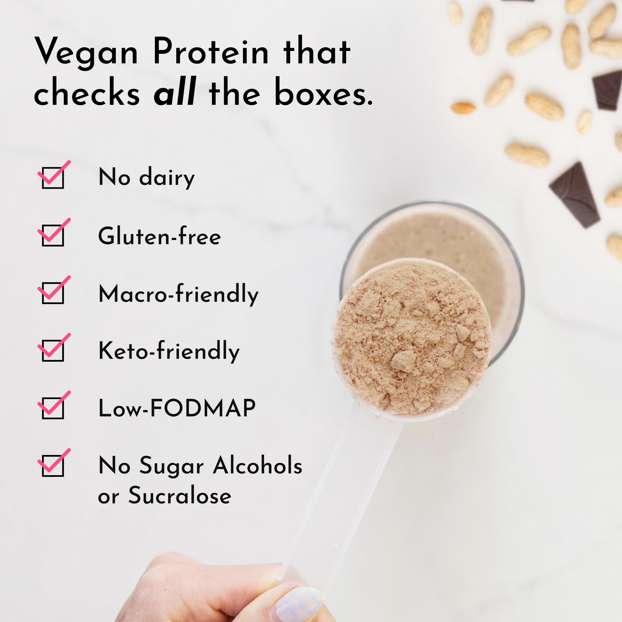 A hand holds a scoop of protein powder above a glass with a smoothie, surrounded by peanuts and chocolate pieces.