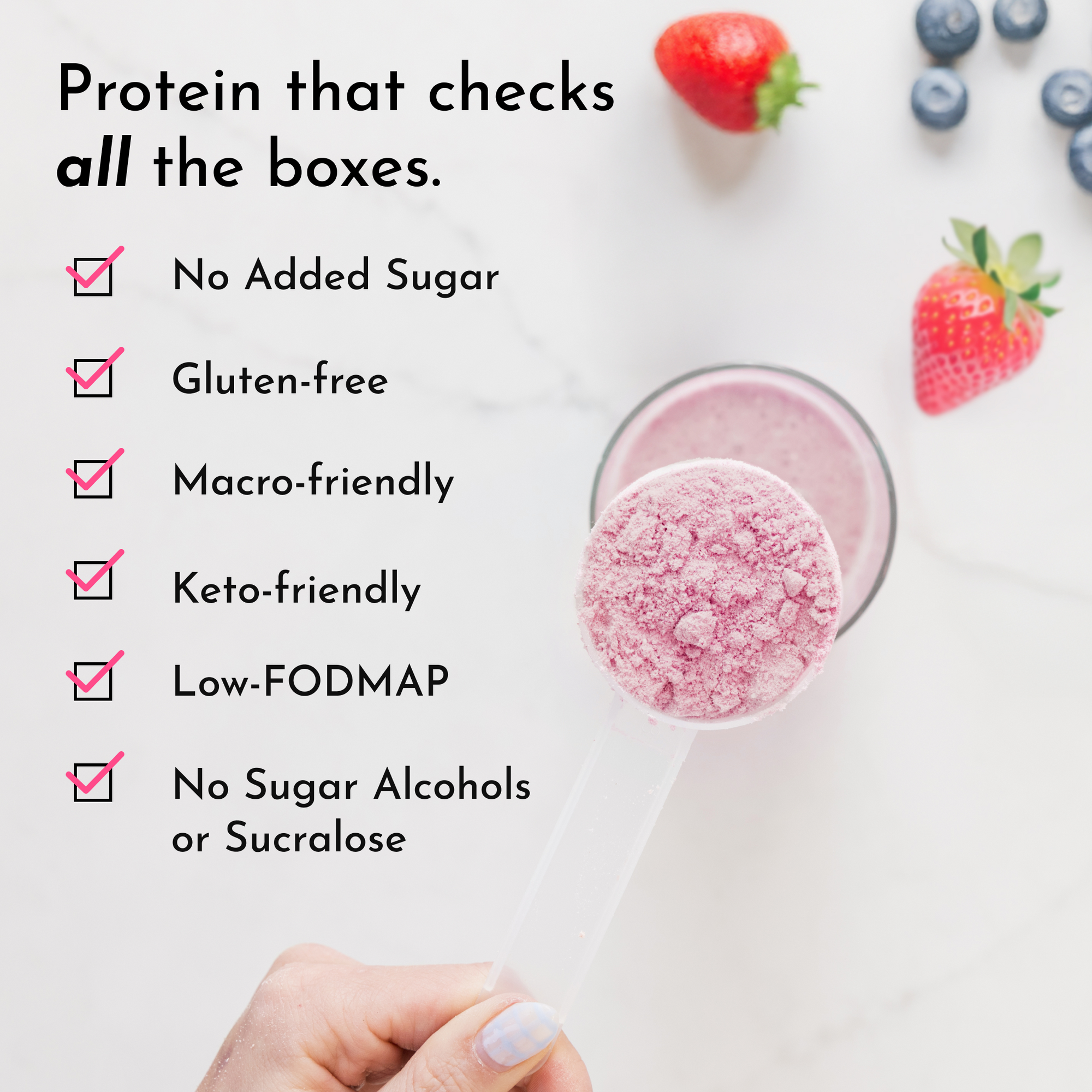 A hand holds a clear scoop of pink protein powder above a glass with a smoothie, surrounded by strawberries and blueberries.