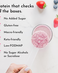 A hand holds a clear scoop of pink protein powder above a glass with a smoothie, surrounded by strawberries and blueberries.