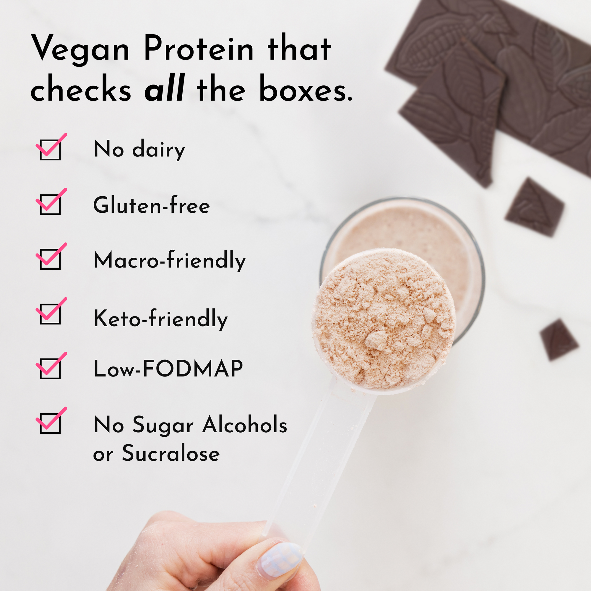 A hand holds a clear scoop of beige protein powder above a glass with a chocolate shake and chocolate pieces in the backgroun