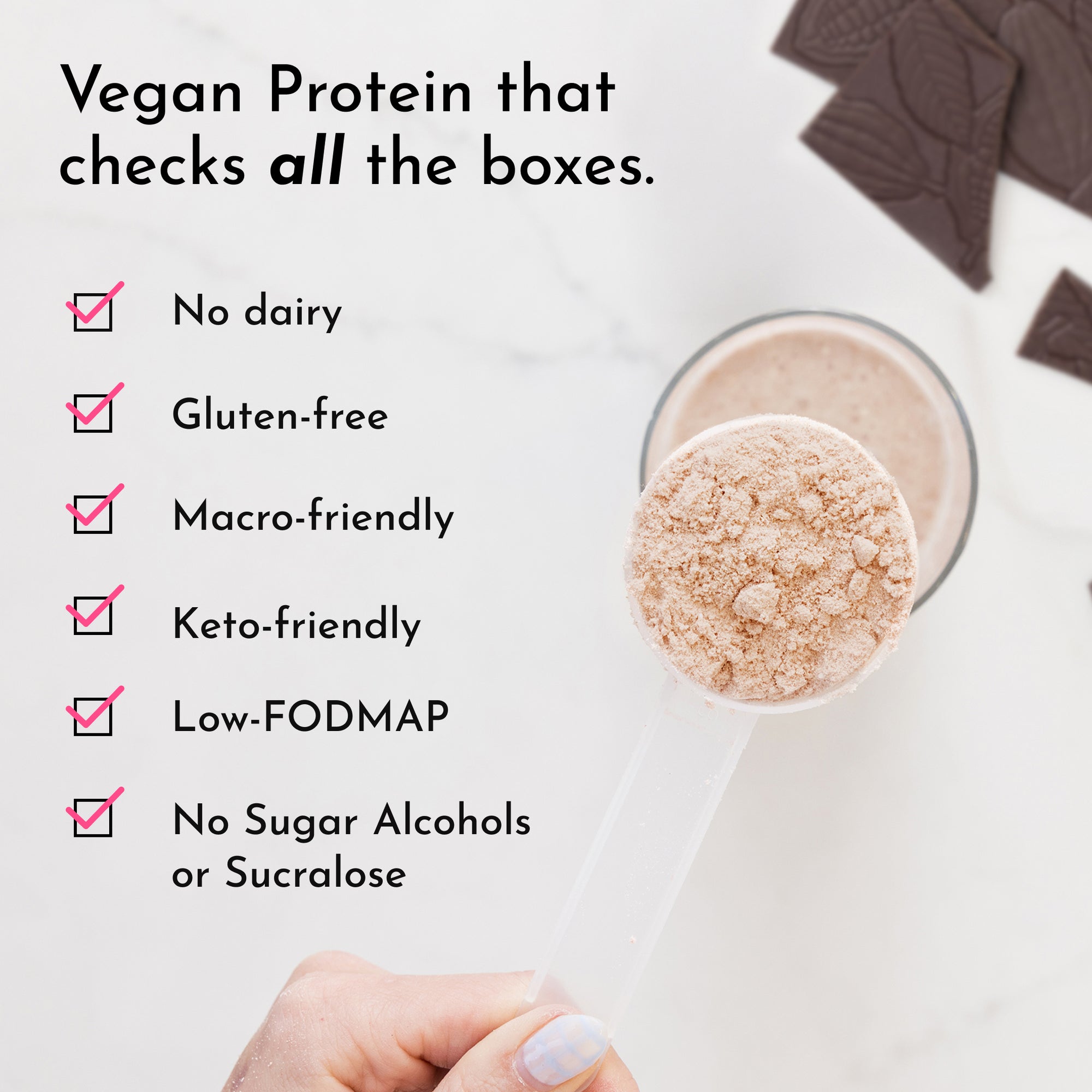 A hand holds a clear scoop of beige protein powder above a glass, with chocolate pieces in the background.