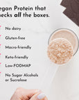 A hand holds a clear scoop of beige protein powder above a glass, with chocolate pieces in the background.