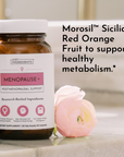 Amber glass bottle of Menopause+ postmenopausal support supplement with white cap, displayed next to a pink flower.