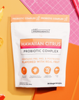 White packet labeled "Hawaiian Citrus Probiotic Complex" with orange gradient banner, surrounded by citrus beverages and pink