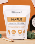 White pouch of Just Ingredients Maple Protein Powder with 22g protein per serving, displayed on orange background with clock 