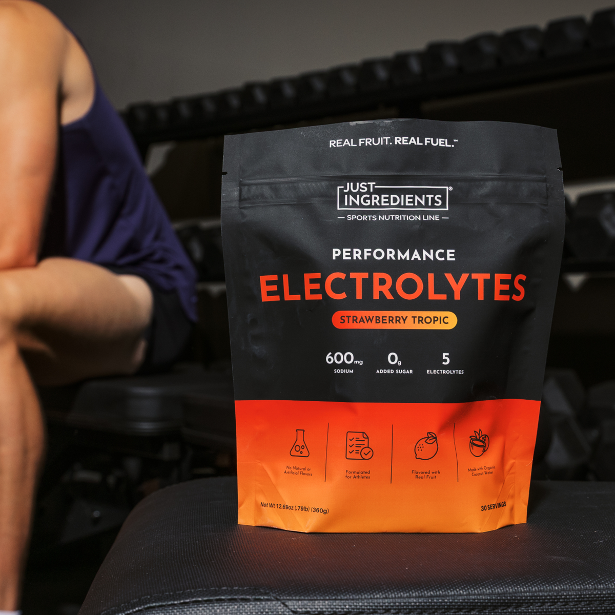 Black and orange pouch of Just Ingredients Performance Electrolytes in Strawberry Tropic flavor held by person in blue tank t