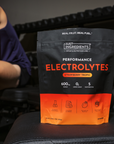 Black and orange pouch of Just Ingredients Performance Electrolytes in Strawberry Tropic flavor held by person in blue tank t