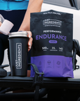 Person holding black tumbler and purple grape endurance drink powder packet with nutrition information displayed.