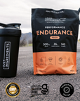Black and orange pouch of peach endurance drink powder next to black shaker bottle on asphalt road.