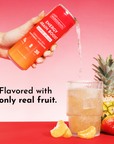 A hand pours a drink from a can into a glass, surrounded by fruit and a pineapple on a colorful background.