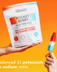 White Rocket Berry Electrolytes powder packet held in one hand with a red, white, and blue popsicle in the other against an o