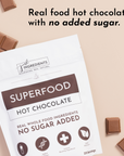 White pouch labeled "Superfood Hot Chocolate" with chocolate squares beside it, featuring text about no added sugar.