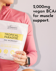 A person in a pink jacket holds a bag of "Tropical Paradise Pre-Workout" with nutritional information visible.