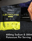 Black and yellow lemon lime electrolyte powder packet on gym floor with person in athletic wear holding it.