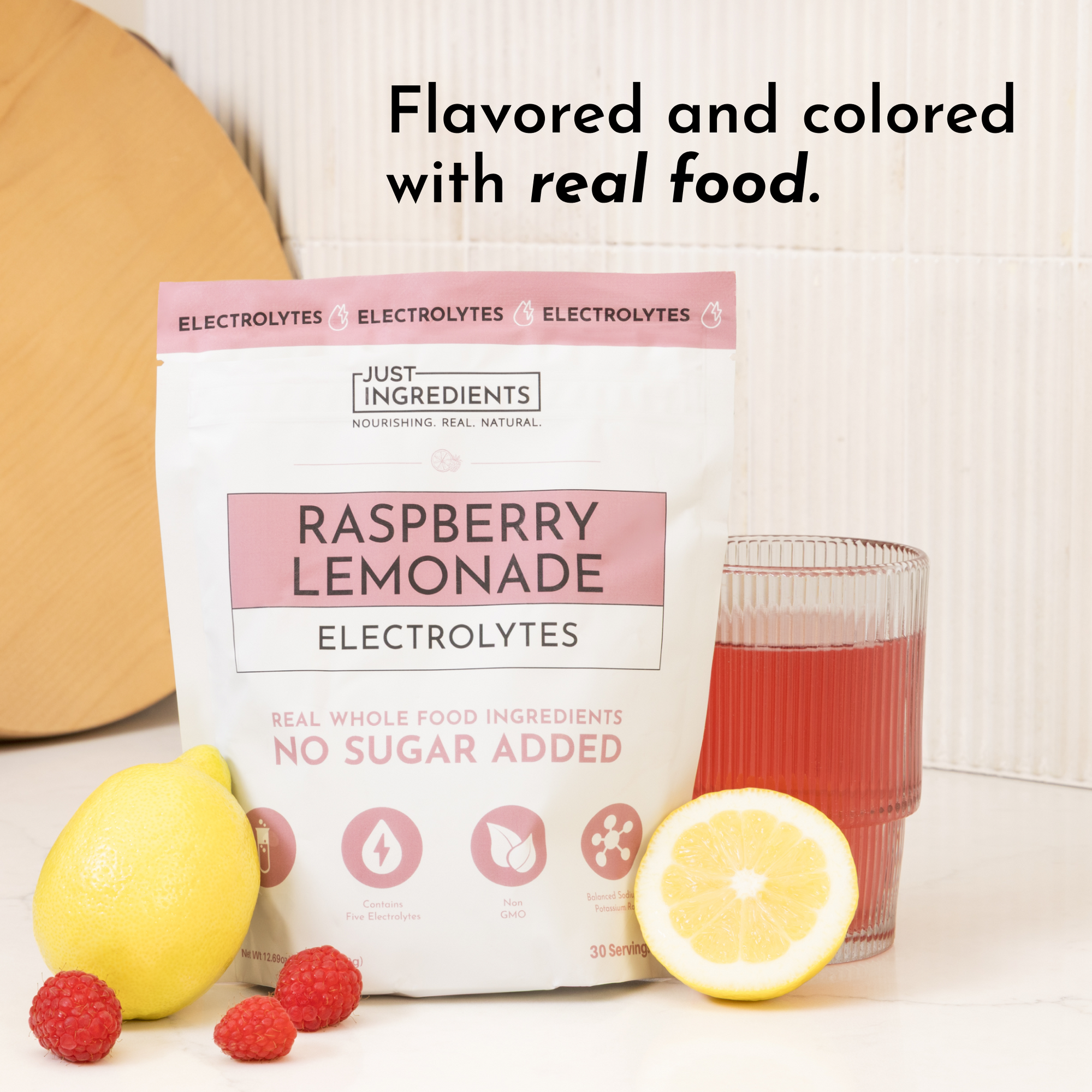 Pouch of Raspberry Lemonade Electrolytes with a glass of red drink, lemon, and raspberries on a white surface.