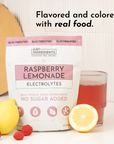 Pouch of Raspberry Lemonade Electrolytes with a glass of red drink, lemon, and raspberries on a white surface.