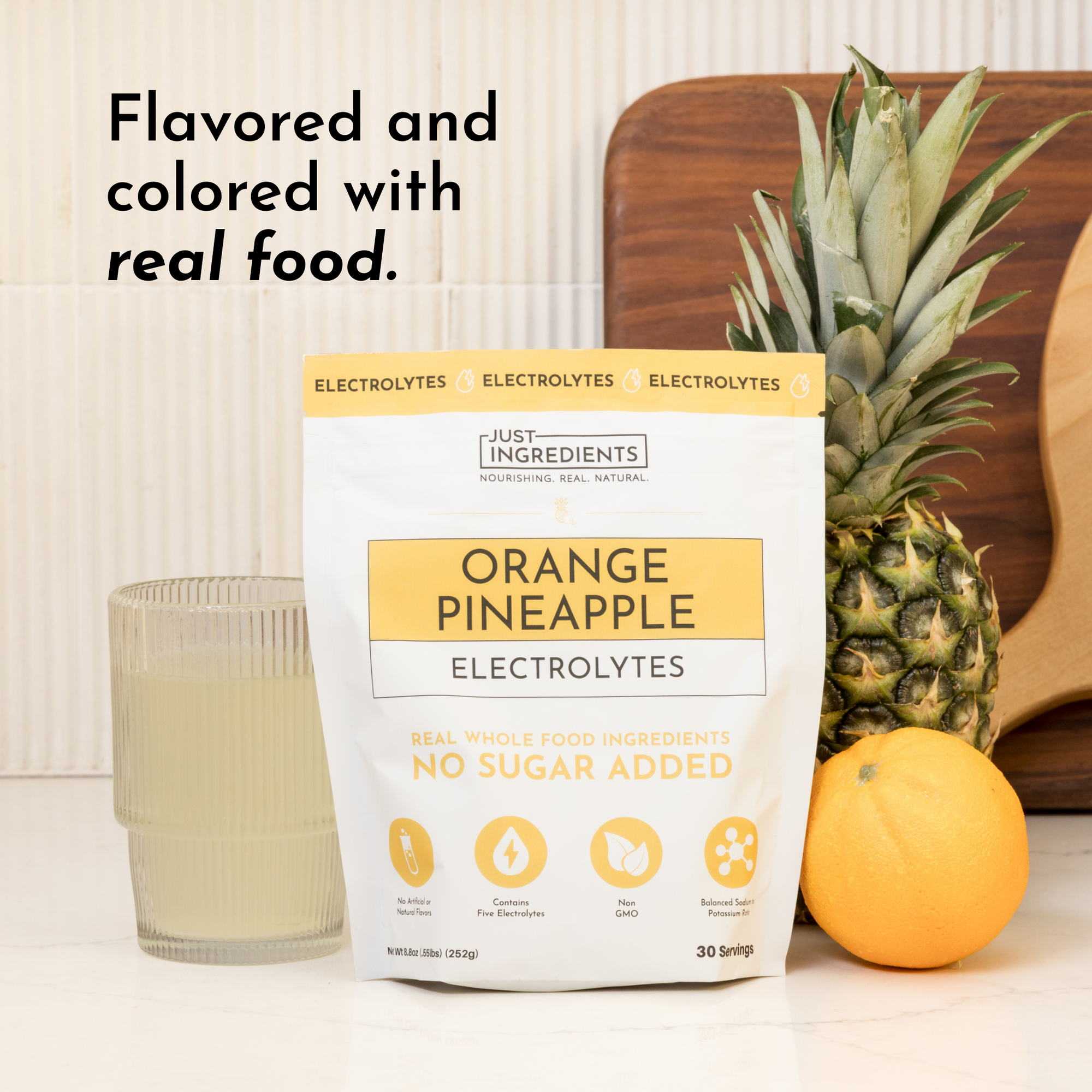 A package of Orange Pineapple Electrolytes next to a glass of liquid, a pineapple, and an orange on a countertop.