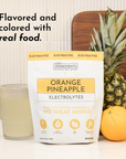 A package of Orange Pineapple Electrolytes next to a glass of liquid, a pineapple, and an orange on a countertop.