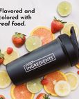 Black shaker bottle with "Just Ingredients" label surrounded by fresh fruits including strawberries, lemons, and oranges.