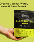 Hand holding black and yellow pouch of lemon lime electrolyte powder against yellow background.