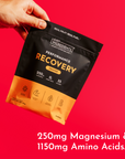 Hand holding black and orange pouch of orange-flavored performance recovery drink against red background.