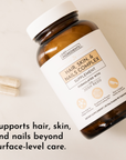 Brown bottle of hair, skin, and nails supplement with two capsules beside it on a light surface.