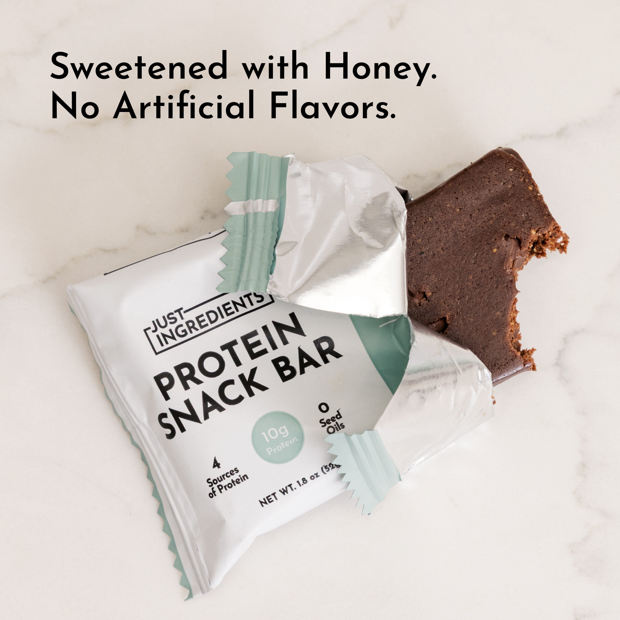 A partially unwrapped protein snack bar with a bite taken out, placed next to its packaging on a marble surface.