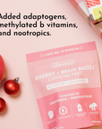 Pink pouch of "Energy + Brain Boost" raspberry lemonade supplement with decorative ornaments and gift wrapping in the backgro